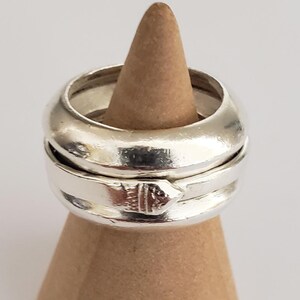 May include: Two silver rings, one thick and rounded, the other thinner with a decorative element, are displayed on a small, brown, wooden cone-shaped stand. The rings are reflective, and the stand has a smooth, natural finish. The background is plain white.