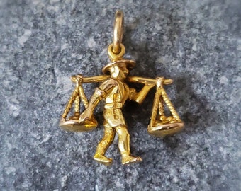 May include: Gold charm of a person carrying two baskets on a pole. The charm is detailed and has a realistic design.