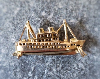May include: A gold-toned ship brooch with a detailed design. The ship has a pointed bow and a single mast with a small sail. The brooch is set on a gray stone background.