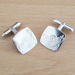 Könnte beinhalten: Ein Paar silberne Manschettenknöpfe mit quadratischem Design. Jeder Manschettenknopf zeigt ein detailliertes, graviertes Blumenmuster.
