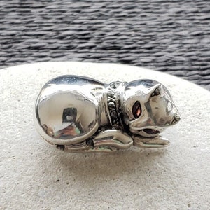 May include: A silver cat charm, lying down with its head turned to the side. The cat is wearing a black and white beaded collar and has red eyes.