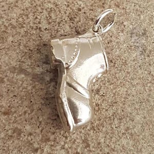 Könnte beinhalten: Ein polierter silberfarbener Metallanhänger in Form eines traditionellen Holzschuhs. Der Anhänger verfügt über einen kleinen runden Ring an der Oberseite zur Befestigung und weist detaillierte Linien auf, die Struktur und Nähte darstellen.