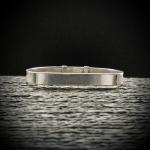 May include: A silver bangle bracelet with a simple, rectangular design. The bracelet is made of polished metal and has a smooth, shiny finish.