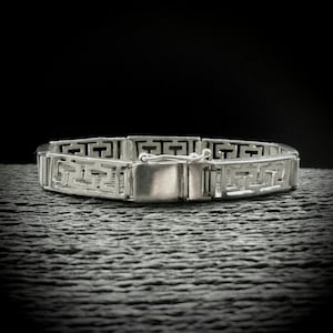 May include: A silver bracelet featuring a geometric design. The bracelet is composed of rectangular links, each displaying a repeating Greek key pattern. The clasp is a simple, rectangular box. It is presented on a textured, dark grey surface.