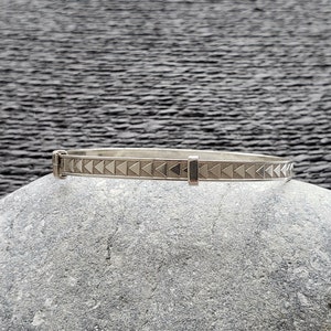 May include: A silver bangle bracelet with a geometric pattern of triangles. The bracelet is on a gray stone surface.
