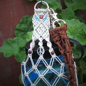 May include: A macrame water bottle holder made of off-white cord featuring turquoise and brown beads, a silver peace sign charm, and a brown suede leather strap attached with a silver metal swivel clasp, secured around a blue plastic water bottle.