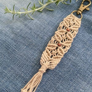 May include: Beige macrame keychain with a bronze-colored clasp and two small wooden beads. The woven design features a tassel at the bottom. The keychain is shown on a blue denim background.
