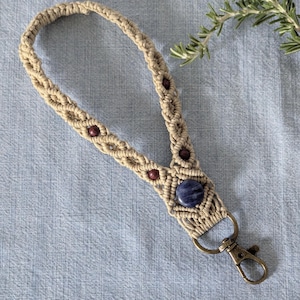 May include: A beige macrame wristlet keychain featuring a braided cord design with small dark red beads and a larger round blue stone accent. The wristlet is attached to a bronze-toned metal lobster clasp for securing keys or other small items.