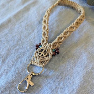 May include: Macrame keychain with a tan woven design, featuring a silver peace sign charm, wooden beads, and a silver clasp. The keychain is handmade and has a bohemian style.