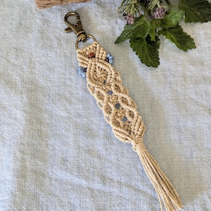 May include: A beige macrame keychain with a bronze-colored clasp and small blue and brown beads. The keychain has a woven design and a tassel at the bottom. The background is a light blue fabric.