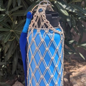 May include: A bright blue water bottle encased in a beige macrame net holder. The holder features a metal ring at the top, a small blue stone accent, and a thick, solid blue fabric strap attached to the side. The bottle has a black lid.