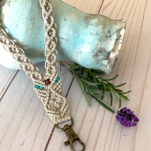 May include: A beige macrame lanyard with turquoise and brown beads. The lanyard has a bronze-colored clasp. The background includes a light blue ceramic vase, sprigs of rosemary, and a sprig of lavender.