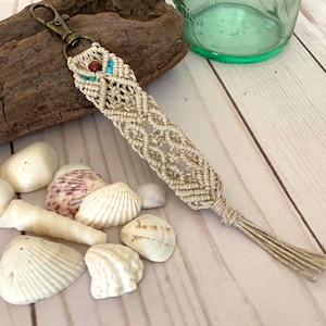 May include: Beige macrame keychain with a bronze-colored clasp and decorative beads. The keychain features a woven design with a tassel at the end. The beads are turquoise and brown. Seashells and driftwood are in the background.