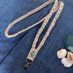 May include: A beige macrame lanyard with a small turquoise turtle charm and gold beads. The lanyard has a metal clasp and is displayed on a blue denim surface. Decorative seashells are in the background.