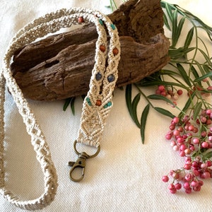 May include: A beige macrame lanyard featuring intricate knotted patterns, accented with small round wooden beads and blue stone beads. The lanyard includes a bronze-toned metal lobster clasp at the bottom for attaching items securely.