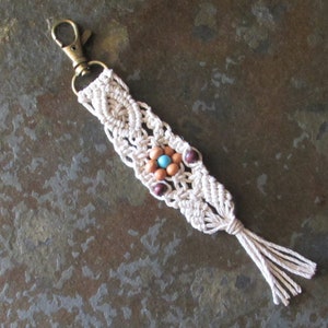 May include: A macrame keychain featuring off-white cotton cord knotted in a diamond pattern, accented with a central cluster of light brown wooden beads, a single bright turquoise bead, and two dark purple beads, finished with a brass lobster clasp.