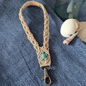 May include: A beige macrame keychain with a turquoise turtle charm and a bronze-colored clasp. The keychain features a woven design with wooden bead accents. The background includes seashells and denim fabric.