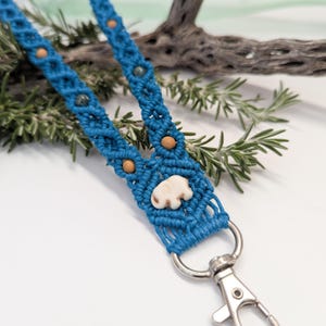 May include: A handmade blue macrame lanyard with a silver-colored clasp. The lanyard features wooden beads and a small, off-white elephant charm. The background includes greenery and a piece of driftwood.