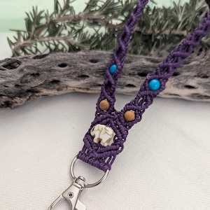 May include: A handcrafted purple macrame lanyard with turquoise and wooden beads. The lanyard features a small, off-white elephant charm and a silver-colored clasp. The background includes a piece of driftwood and greenery.