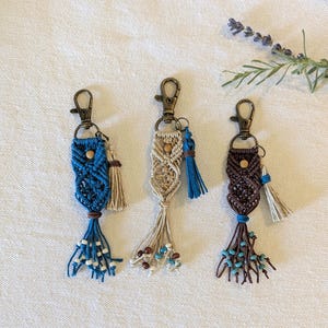 May include: Three macrame keychains featuring bronze-toned lobster clasps, intricate knotted cord patterns in blue, beige, and dark brown, wooden beads, and decorative tassels with small accent beads at the ends of the hanging cord strands.