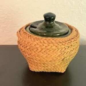May include: A small, round, woven basket with a green ceramic lid. The basket is made of natural materials and has a woven pattern.