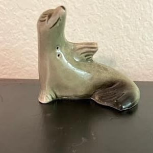 May include: A ceramic figurine of a grey seal with its flippers raised, as if it is waving. The seal is sitting on a black surface.