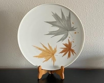 May include: A white ceramic plate with a brown wooden stand. The plate has a design of three stylized leaves in shades of brown and gray.
