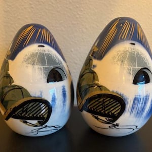 May include: Two egg-shaped ceramic figurines. Each has a white base with a painted design featuring blue, gold, and green accents. The top portion is blue with gold lines. The design includes a black, stylized element resembling a bird or insect.
