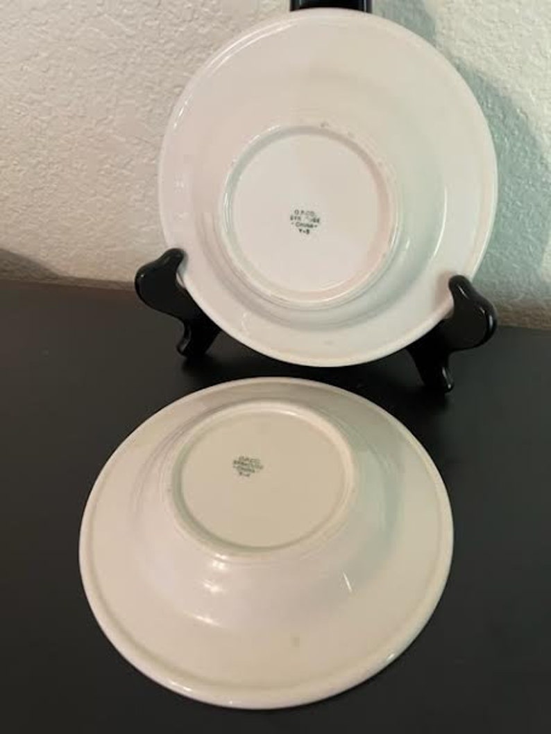 May include: Two white ceramic bowls with a raised rim. The bowls have a small, circular indentation in the center of the bottom. The bowls are marked with the text "Grace  England  China  514".