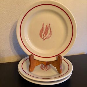 May include: A stack of white ceramic plates with a red rim and a pink floral design in the center. One plate is displayed on a wooden stand. The plates are stacked on a dark surface.