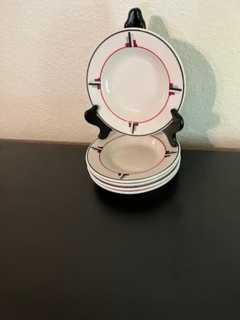 May include: Set of four white ceramic soup bowls with a red, black, and gray geometric design. The bowls are stacked on a black stand.