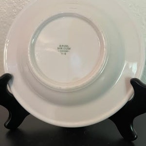 May include: White ceramic saucer with a raised rim. The saucer has a smaller, centered saucer with the text "O.P. & CO. SYRACUSE CHINA Y-8" printed on it.