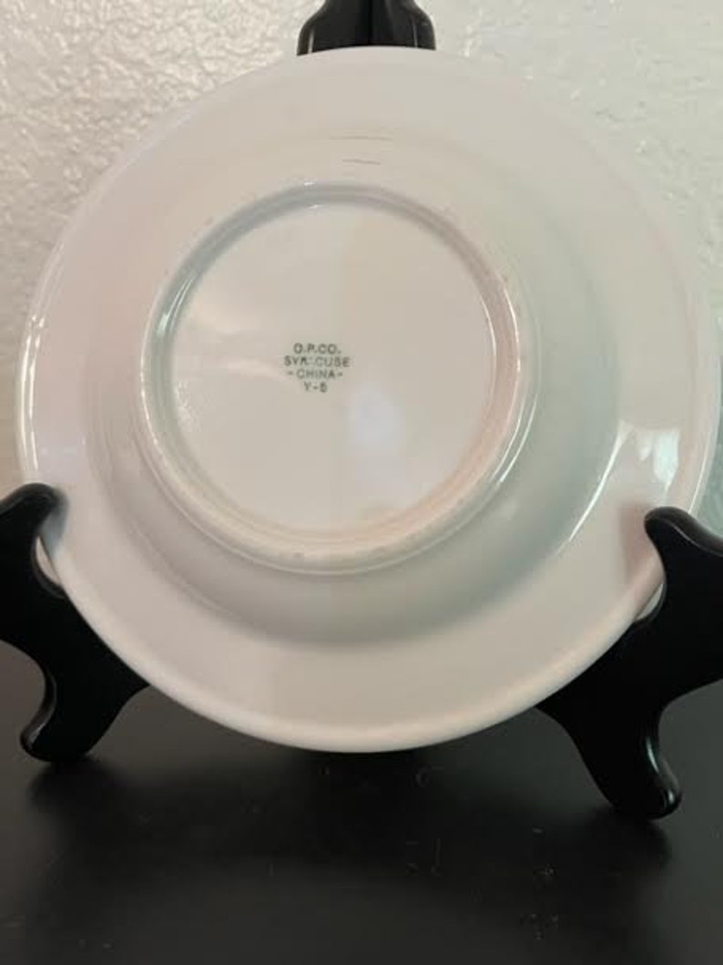 May include: White ceramic saucer with a raised rim. The saucer has a smaller, centered saucer with the text "O.P. & CO. SYRACUSE CHINA Y-8" printed on it.