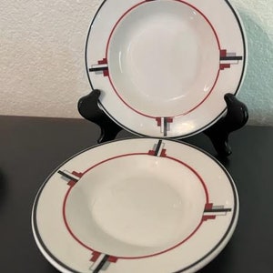 May include: Two white ceramic soup bowls with a geometric design in black, red, and gray. The bowls have a wide rim and a shallow depth.