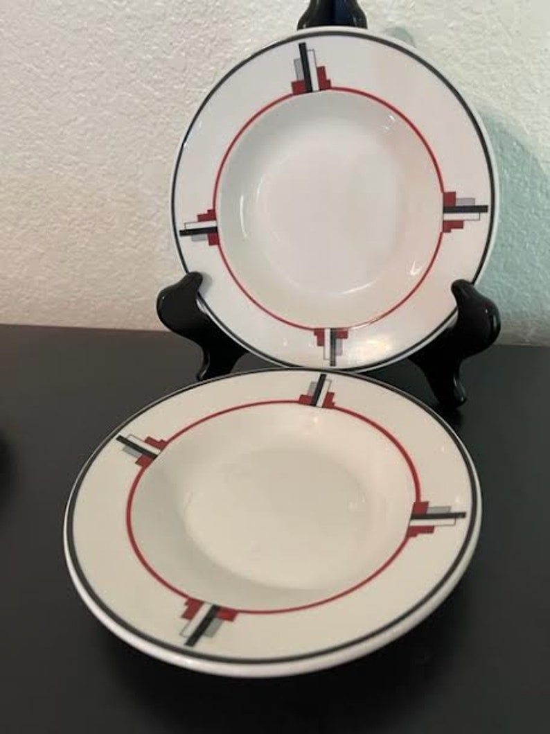 May include: Two white ceramic soup bowls with a geometric design in black, red, and gray. The bowls have a wide rim and a shallow depth.