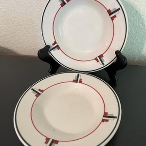 May include: Two white ceramic soup bowls with red, black, and gray geometric designs. The bowls are on a black surface.
