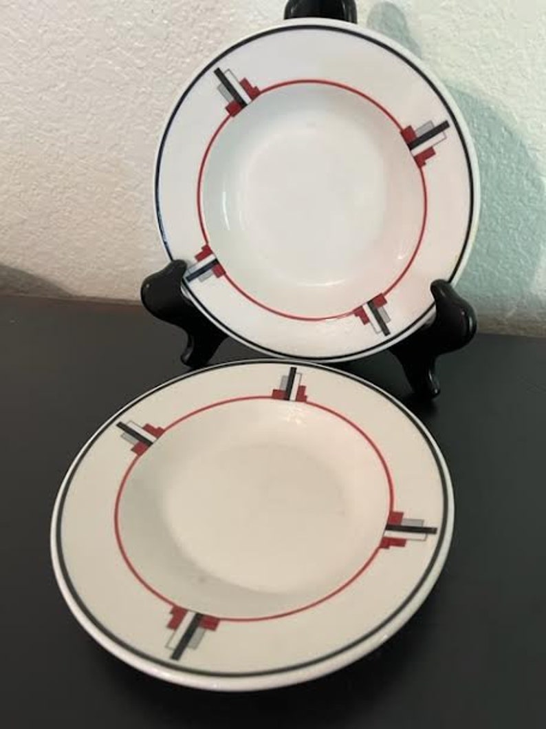 May include: Two white ceramic soup bowls with red, black, and gray geometric designs. The bowls are on a black surface.