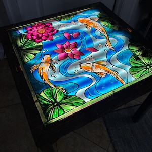 May include: Illuminated table with a stained glass design featuring koi carp, water lilies, and flowing water. The artwork showcases orange fish, pink flowers, and green lily pads against a blue and white background. The table has a dark wooden frame.