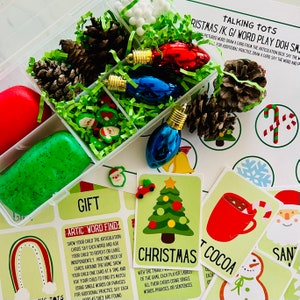 Peut inclure: Un ensemble d'activités de pâte à modeler sur le thème de Noël pour les enfants. L'ensemble comprend un récipient en plastique transparent avec de la pâte à modeler verte et rouge, de petites décorations de Noël, des pommes de pin et un jeu de cartes avec des mots et des images sur le thème de Noël. Les cartes comprennent des mots comme "Noël", "chocolat chaud" et "Père Noël".