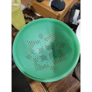 May include: A round, light green plastic colander featuring a series of small, circular drainage holes arranged in a geometric pattern across the interior base and sides, designed for straining liquids from food items during preparation.