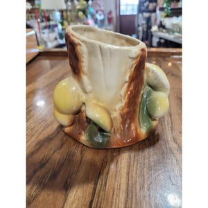 Puede incluir: Una maceta de cerámica con forma de tocón de árbol con interior color crema y exterior marrón. La base presenta detalles de hongos moldeados en amarillo y verde adheridos a los lados del tocón, creando una apariencia orgánica y texturizada.