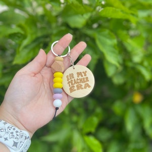 May include: A keychain with a wooden pendant that reads "IN MY TEACHER ERA" in brown lettering. The keychain has a series of colorful beads in yellow, gray, lavender, and white. The keychain is held in a hand against a backdrop of green foliage.