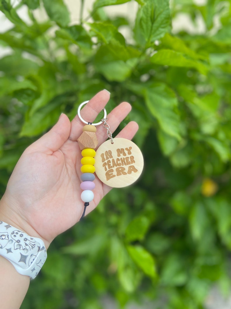 May include: A keychain with a wooden pendant that reads "IN MY TEACHER ERA" in brown lettering. The keychain has a series of colorful beads in yellow, gray, lavender, and white. The keychain is held in a hand against a backdrop of green foliage.
