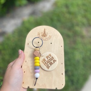 May include: A wooden keychain with a beaded design in yellow, gray, purple, and white. The keychain has a wooden tag that says "IN MY TEACHER ERA". The keychain is attached to a wooden display board.