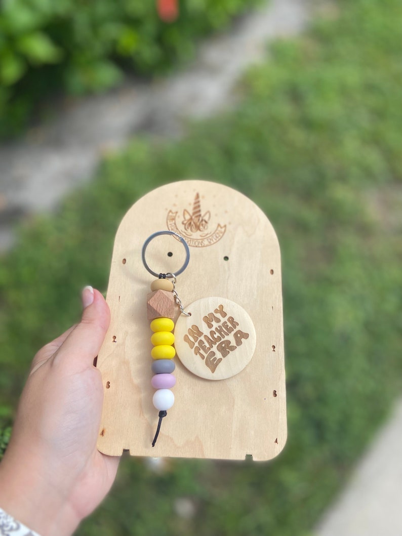 May include: A wooden keychain with a beaded design in yellow, gray, purple, and white. The keychain has a wooden tag that says "IN MY TEACHER ERA". The keychain is attached to a wooden display board.