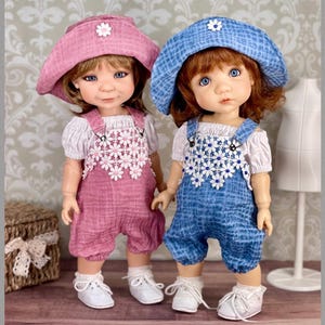 May include: Two dolls wearing matching outfits. One doll is in a pink outfit and the other is in blue. Each doll wears a matching hat with a white flower, a white blouse, and white shoes. The dolls are standing on a wooden surface.