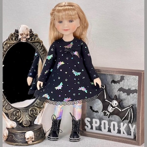 May include: A doll in a black dress with bat and star patterns, iridescent leggings, and black boots. The dress has a lace trim. A decorative mirror and a sign that says "SPOOKY" are in the background.