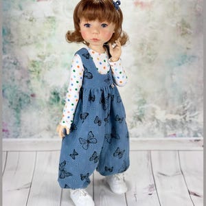 Puede incluir: Una muñeca con un vestido de peto azul con una camisa blanca de lunares y un estampado de mariposas. La muñeca tiene el pelo castaño y los ojos azules.