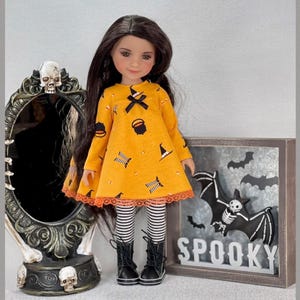 May include: A doll wearing a Halloween-themed outfit. The doll is dressed in an orange dress with black witch hat and cauldron graphics, paired with striped leggings and black boots. A decorative mirror and a sign that says "Spooky" are in the background.