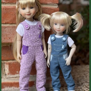 Może przedstawiać: Dwie lalki w ogrodniczkach. Lalka po lewej stronie ma na sobie fioletowe ogrodniczki w białe kropki i białą koszulę. Lalka po prawej stronie ma na sobie niebieskie dżinsowe ogrodniczki z białą koszulą. Obie lalki mają blond włosy i stoją przed ceglaną ścianą.